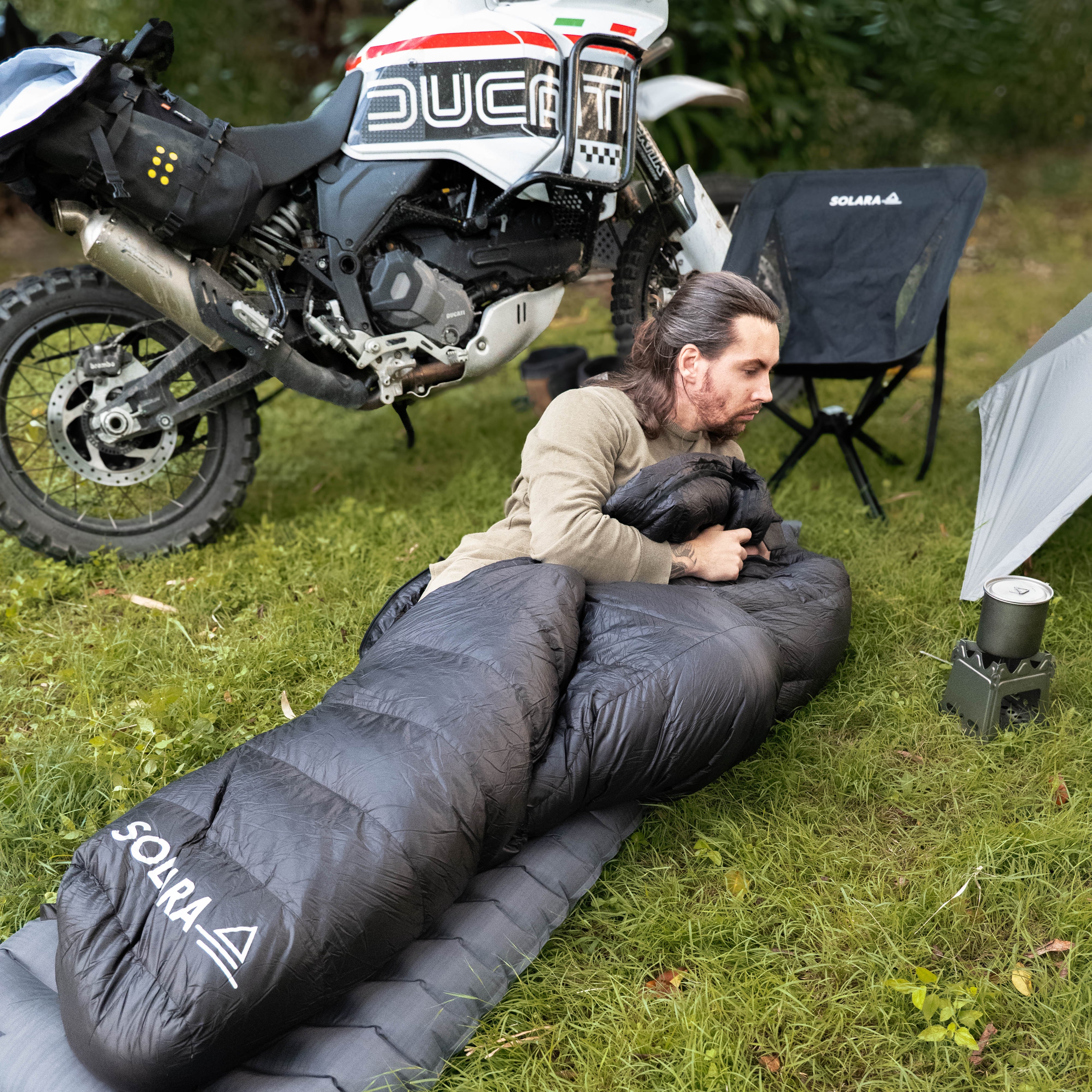 Person lying on an inflatable mattress with a sleeping bag, next to a Ducati motorcycle in a grassy area.