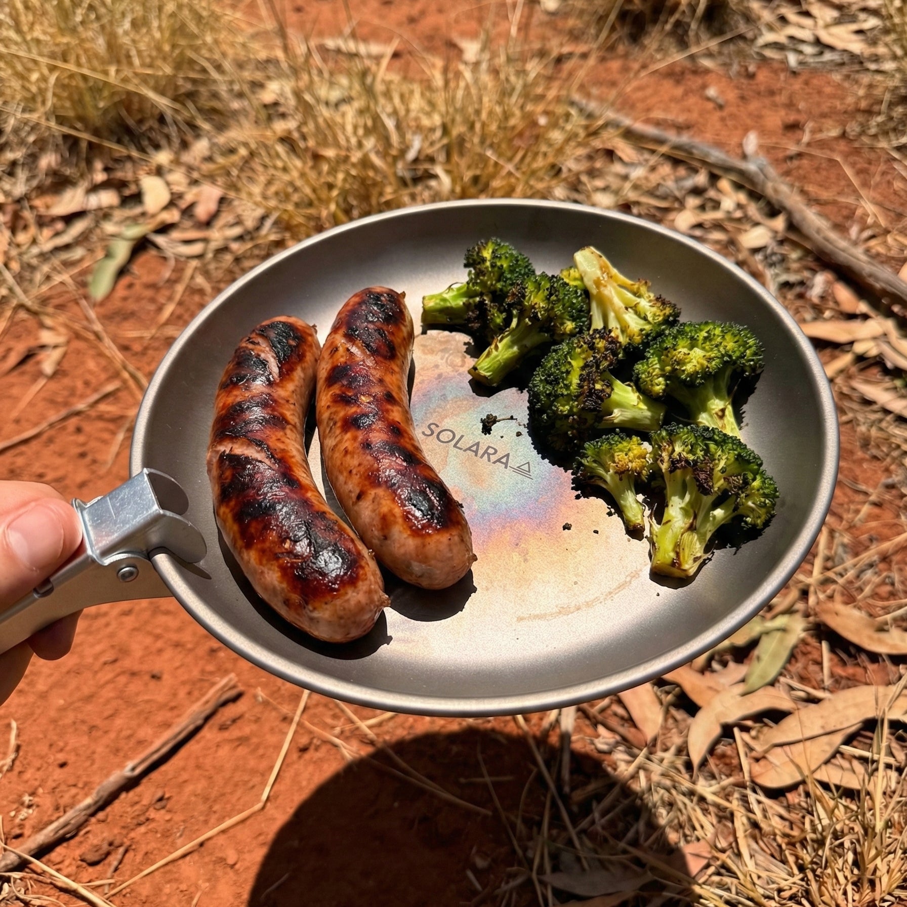 Fried sausages and broccoli in a frying pan held over a red dirt background