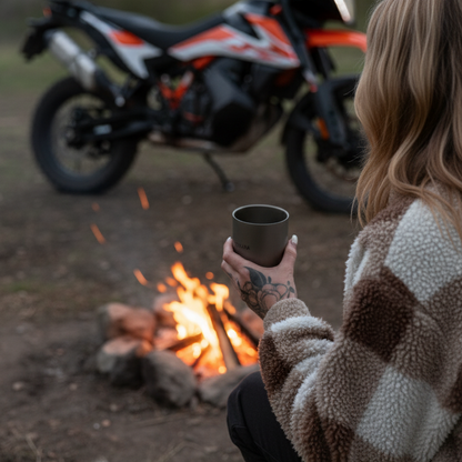 Person holding a mug by a campfire with a motorcycle in the background