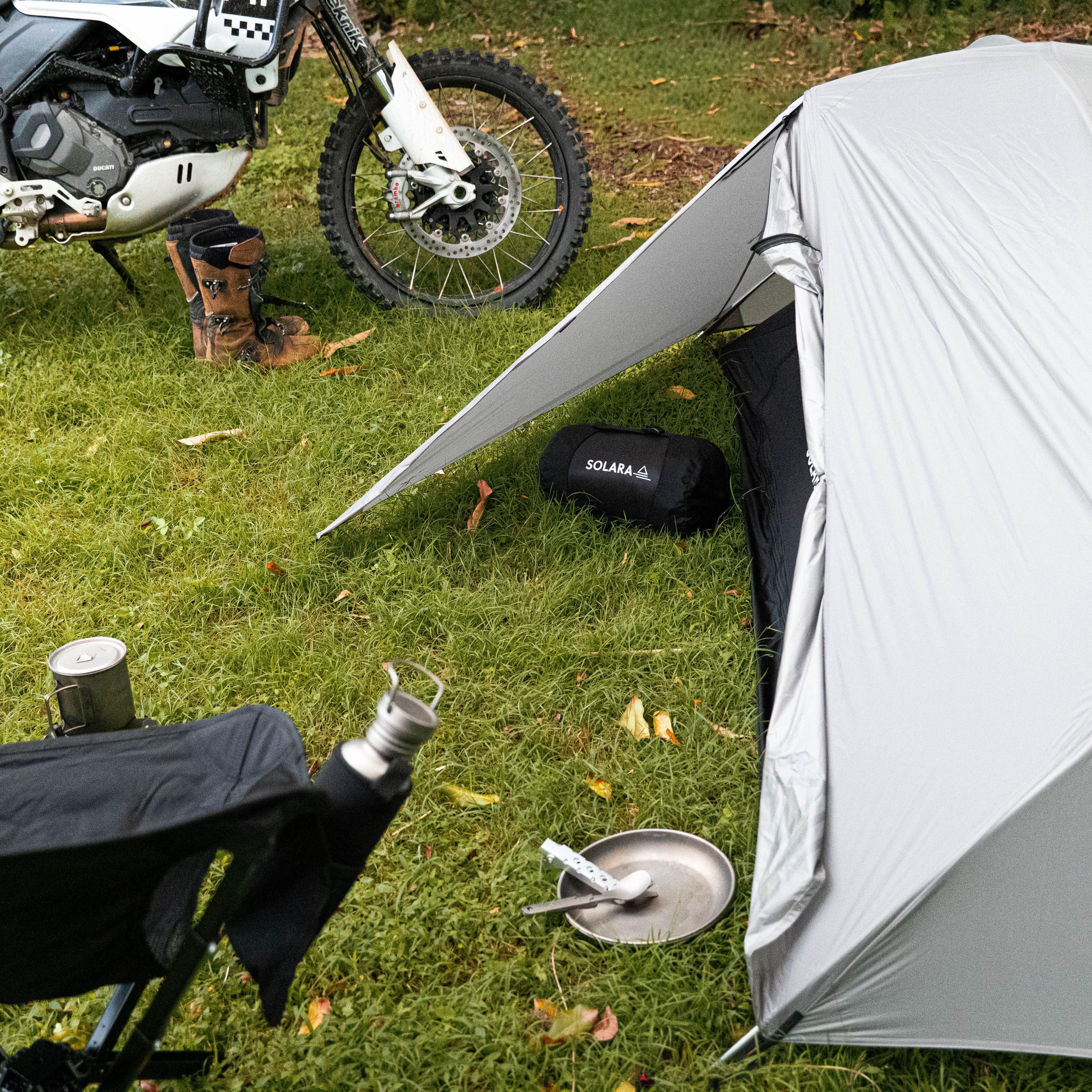 Motorcycle and camping gear including a tent, chair, and camping stove on grass.