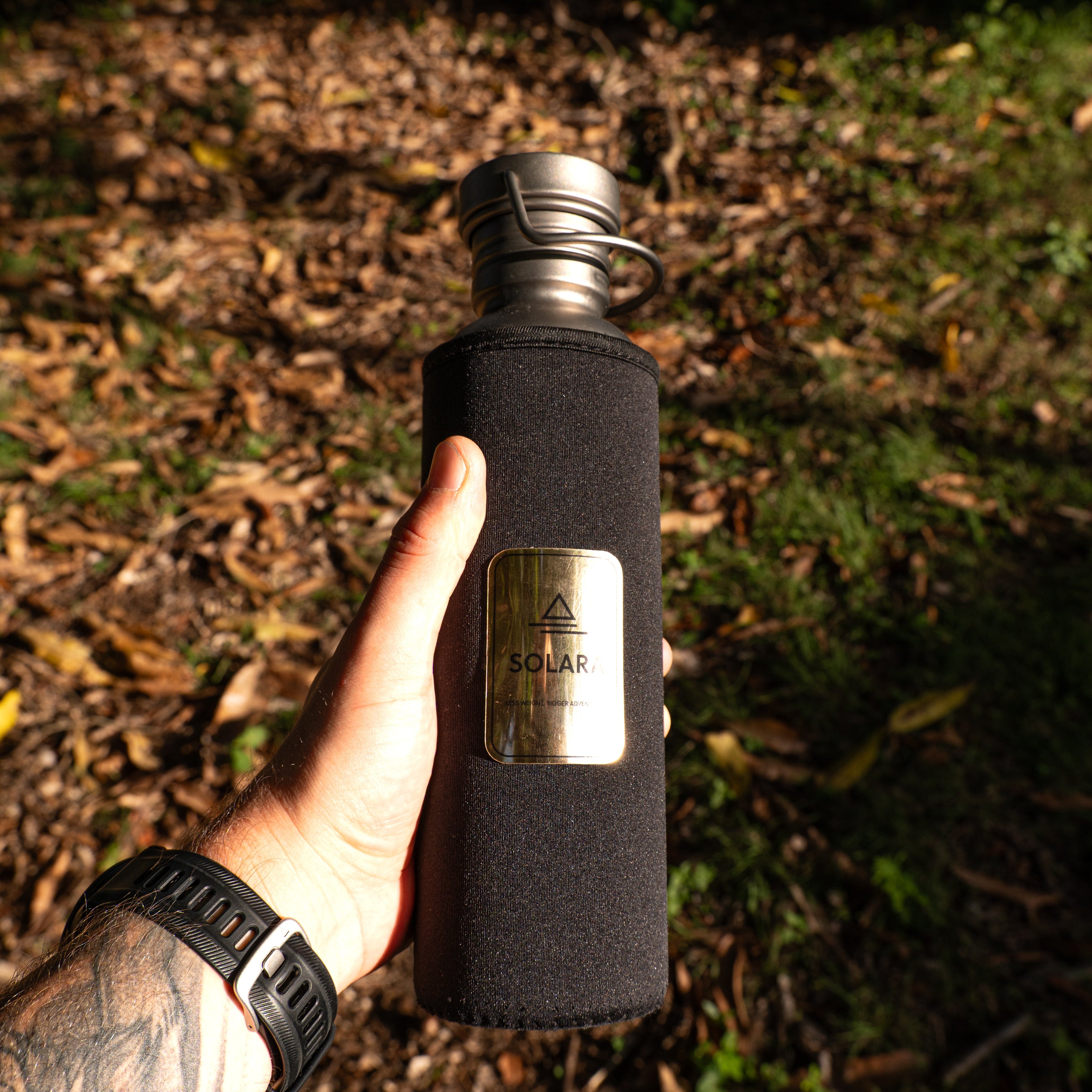 Person holding a black insulated water bottle with a logo in a natural setting